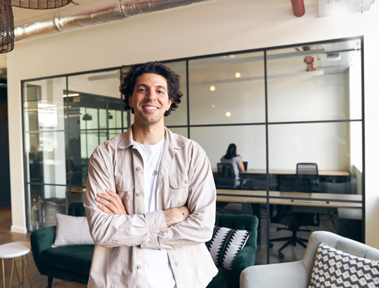 smiling man standing in modern office setting with green couch and workspace in background featuring two people interacting