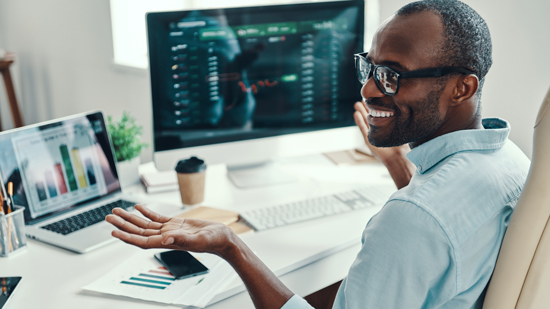 smiling man with glasses at desk with multiple computer screens displaying graphs and data showing performance analytics and trading strategies related to two financial assets