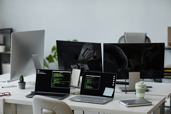 three computers with coding screens on a modern office desk including a cup and plant