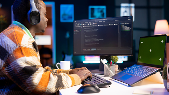 person wearing headphones working on a computer with code on the screen and a laptop nearby focused on multiple tasks related to coding and programming 5 key elements